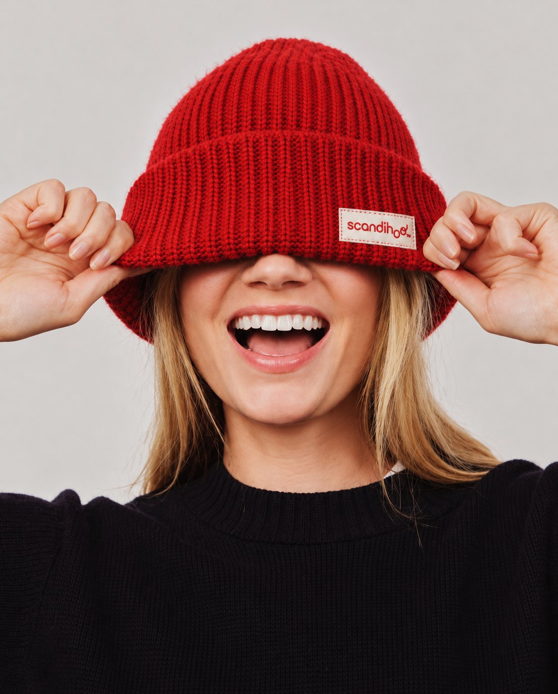 A laughing woman pulling a red knit hat with a Scandihoo label down over her eyes